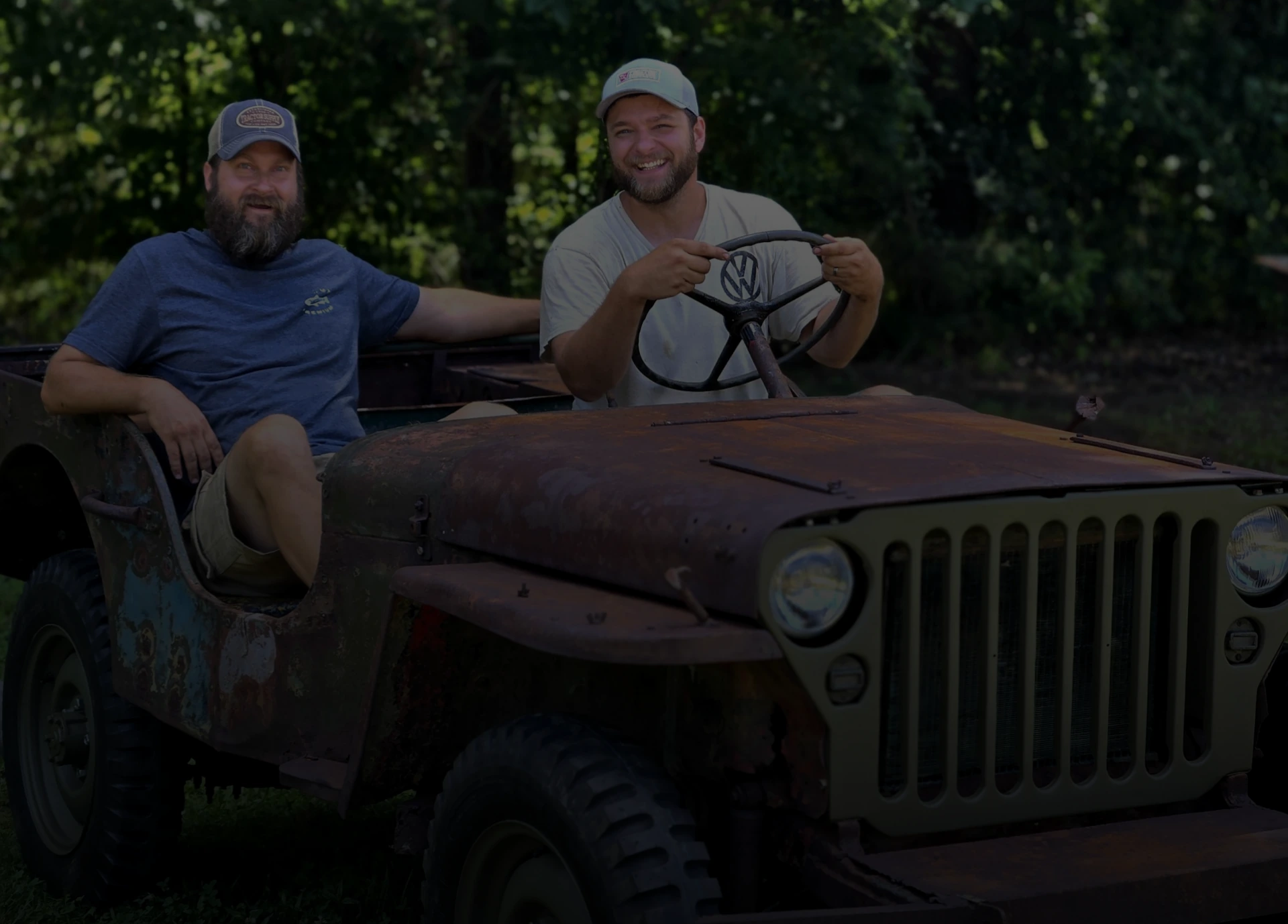 Two men sitting in a rustic vintage Jeep outdoors, one driving and smiling at the camera, with a wooded background.