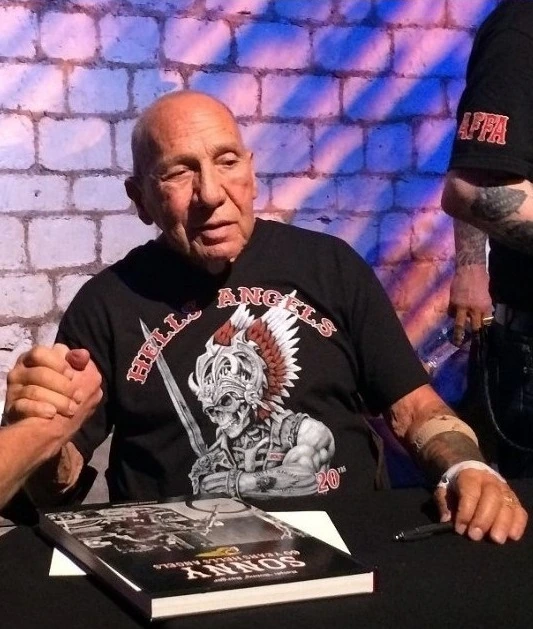 Sonny Barger seated at a table, wearing a Hells Angels shirt, with a book in front of him