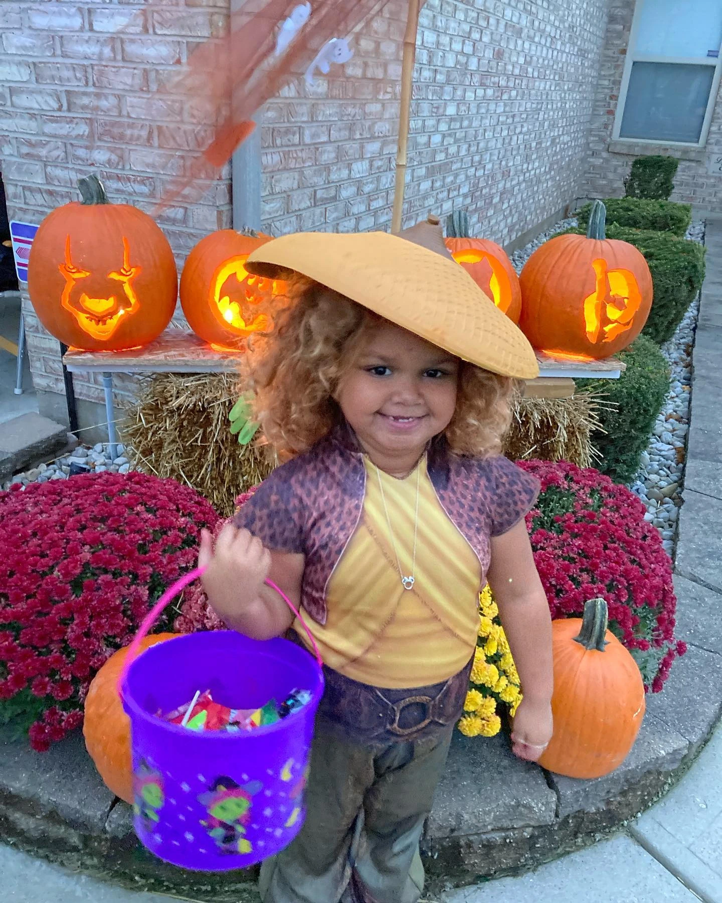 Young child smiling in costume at a pumpkin display, holding a trick-or-treat bucket.